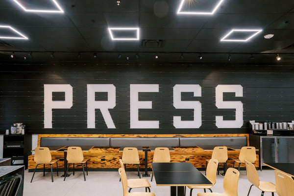 A large PRESS logo made out of custom cut wood slats at our Scottsdale Waterfront location. Moden, sleek wooden chairs and tables with bright white lighting are features of the space. 