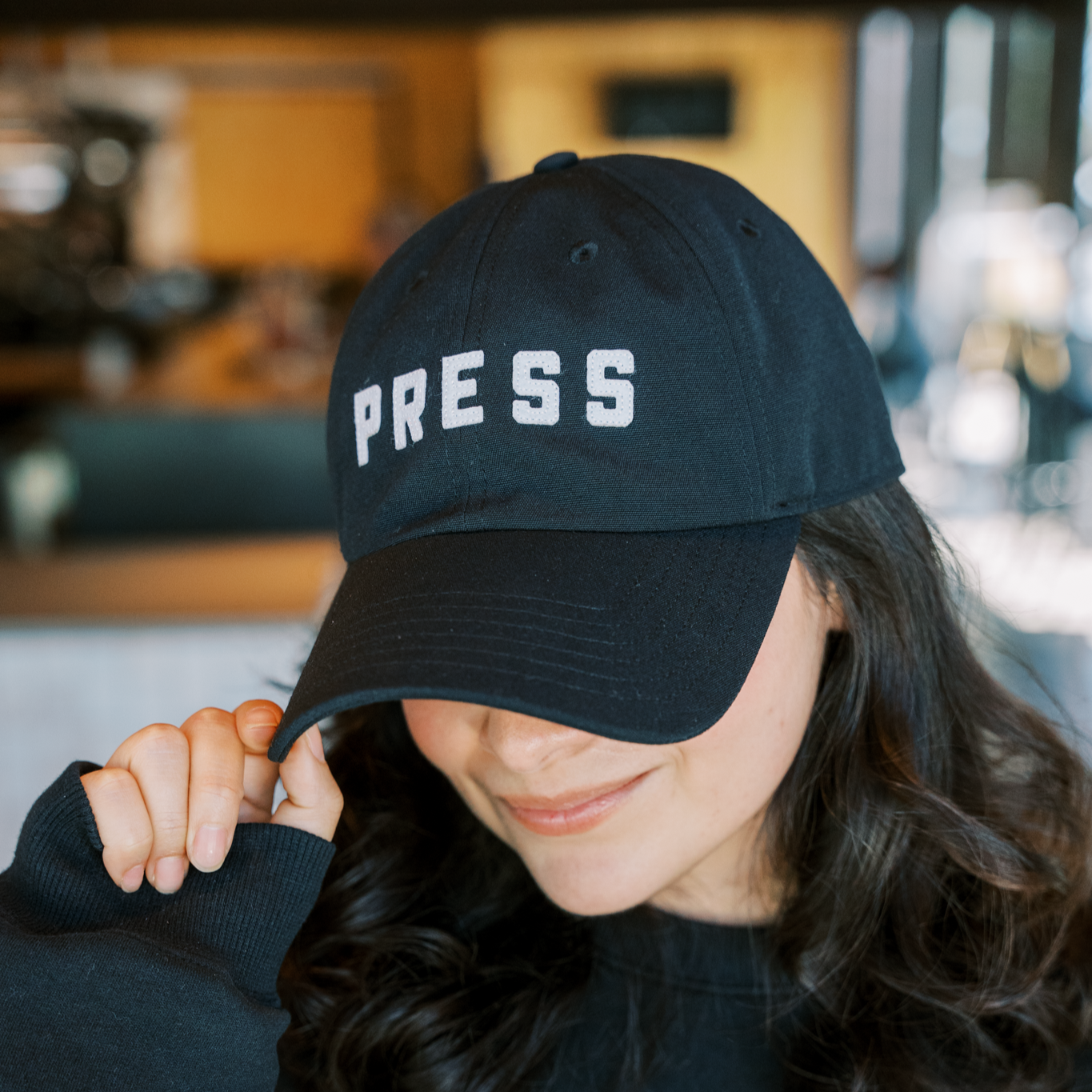 A woman with dark hair wearing a PRESS hat. 