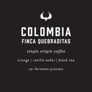 A dark charcoal label with white text reading 'COLOMBIA FINCA QUEBRADITAS' and smaller text 'single origin coffee', 'orange | vanilla wafer | black tea', and 'co-ferment process', with a white winged emblem above the main text.
